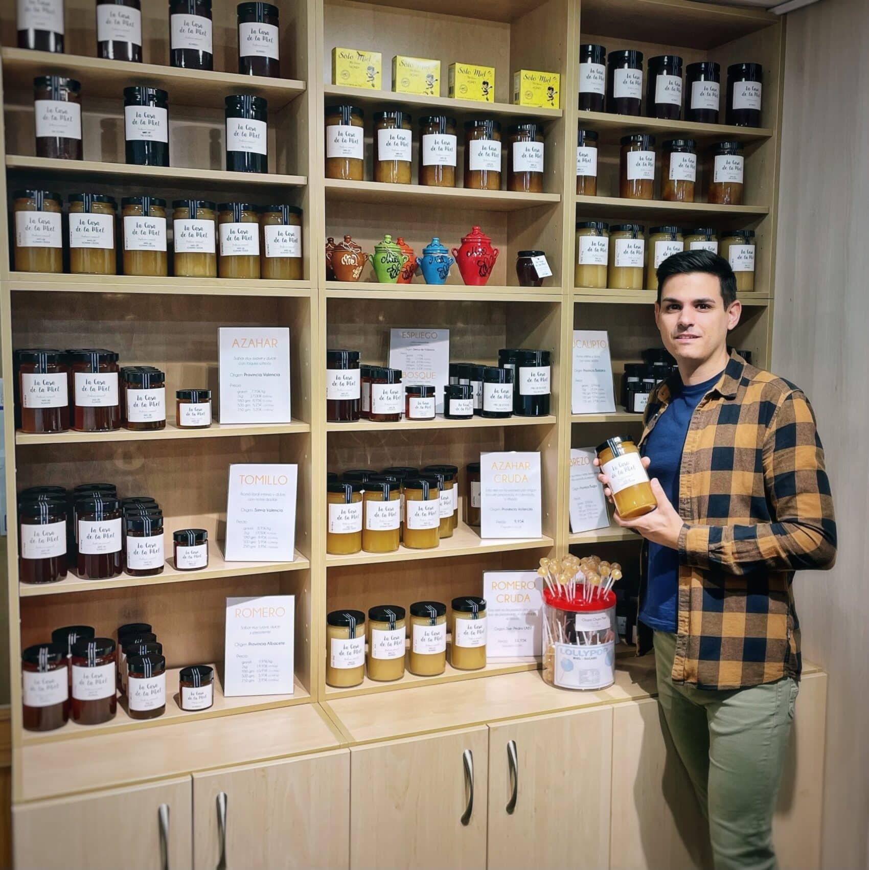 Álvaro con un bote de miel de azahar cruda delante del expositor de La Casa de la Miel donde están todos los tipos de mieles que tenemos y sus distintos formatos.